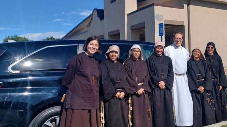 Six women in religious garments and one Catholic priest in religious garments smiling for the camera in front of a vehicle