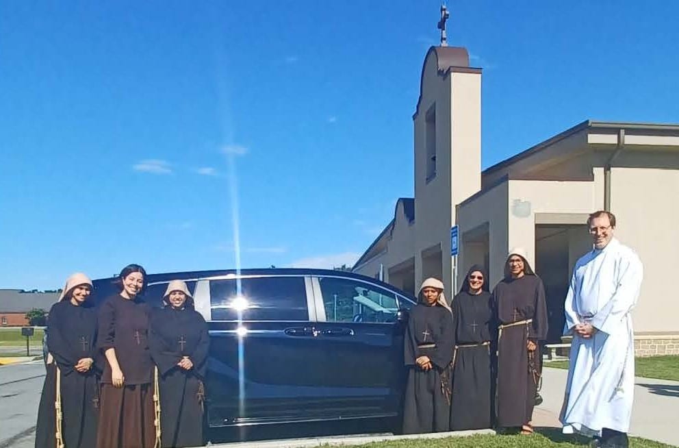 Six women in religious garments and one Catholic priest in religious garments smile for the camera on either side of a vehicle