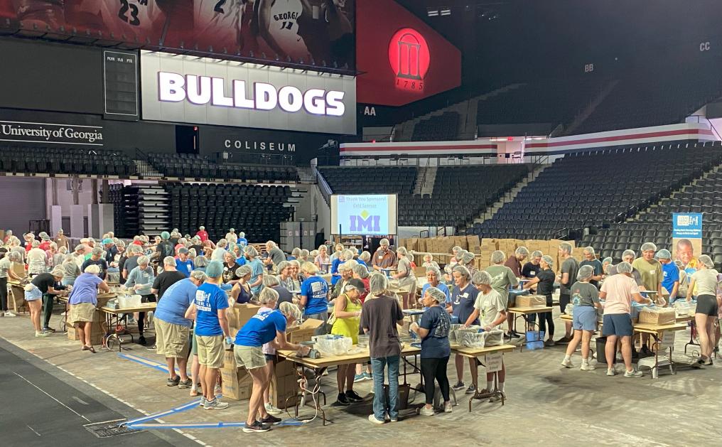 Volunteers Working Together At The Athens Feed My Starving Children Event Around 100 people of all ages work to pack meals as they surround various tables scattered throughout a sports arena.