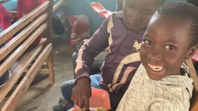 Children sitting on an indoor bench smiling as they share a meal