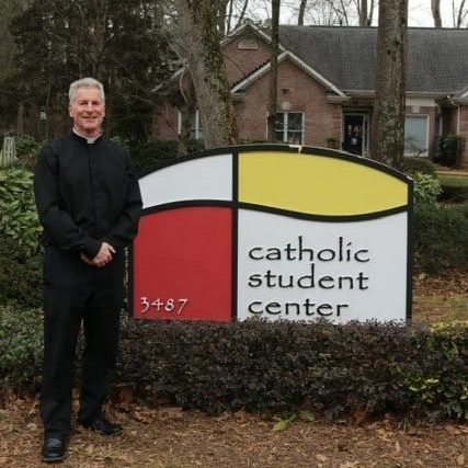 A priest poses in front of a sign that says Catholic Student Center