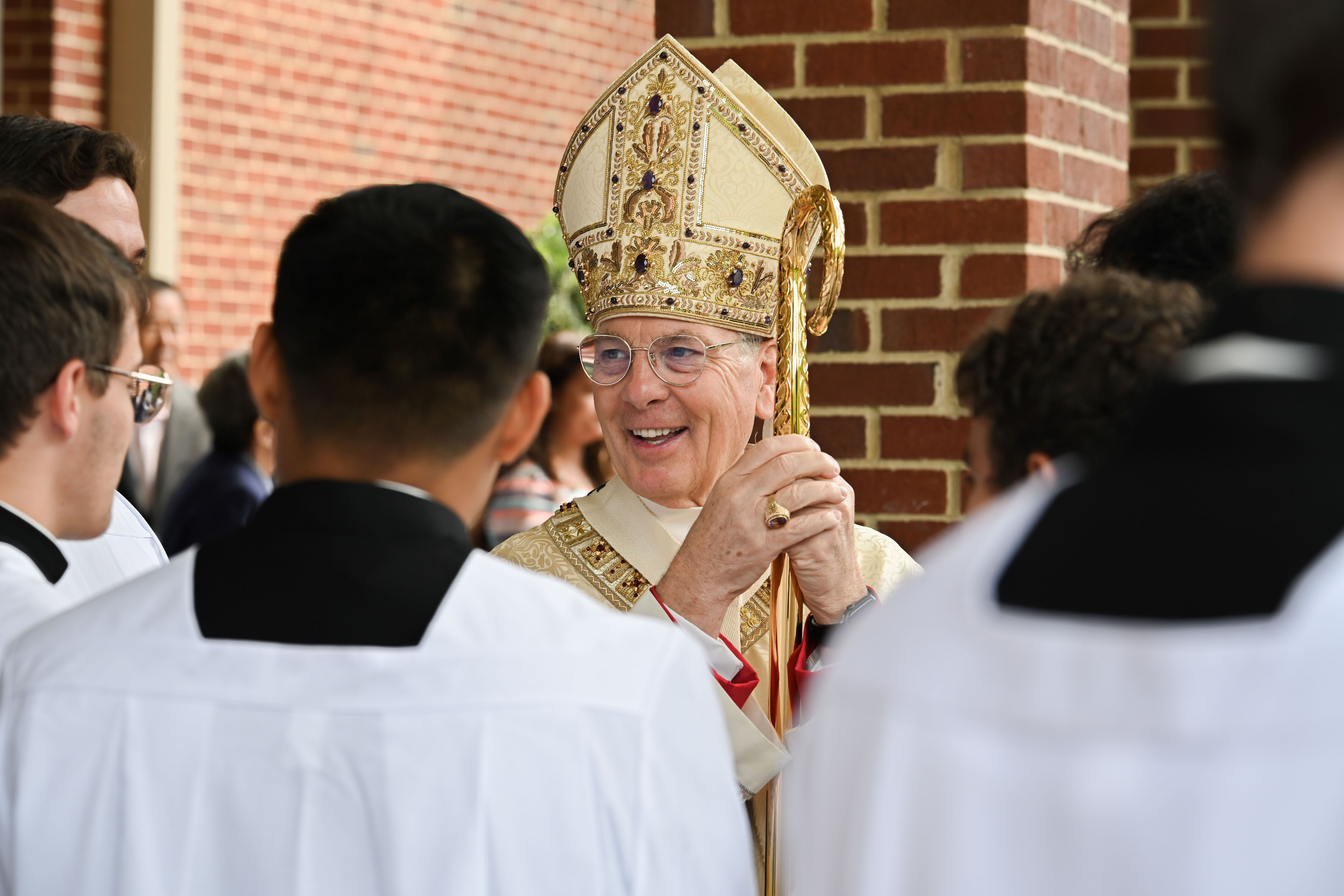 A bishop in celebratory attire smiles talking to men in clergy attire