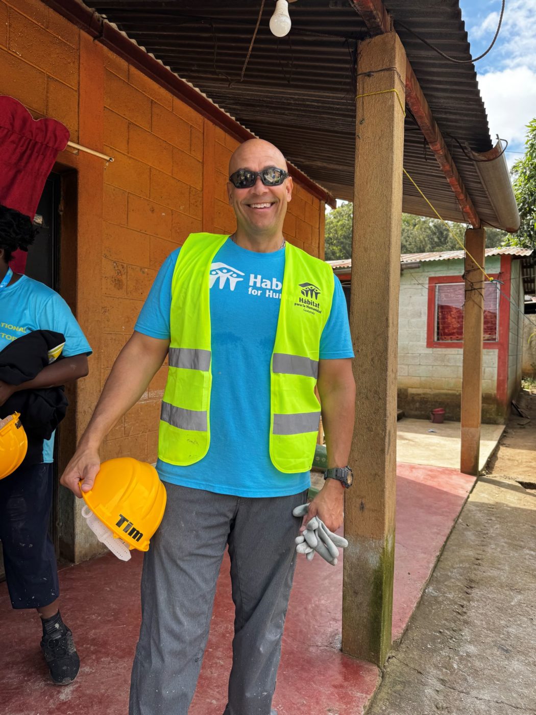 A man in a construction vest, holds a helmet and gloves while smiling in front of a construction site