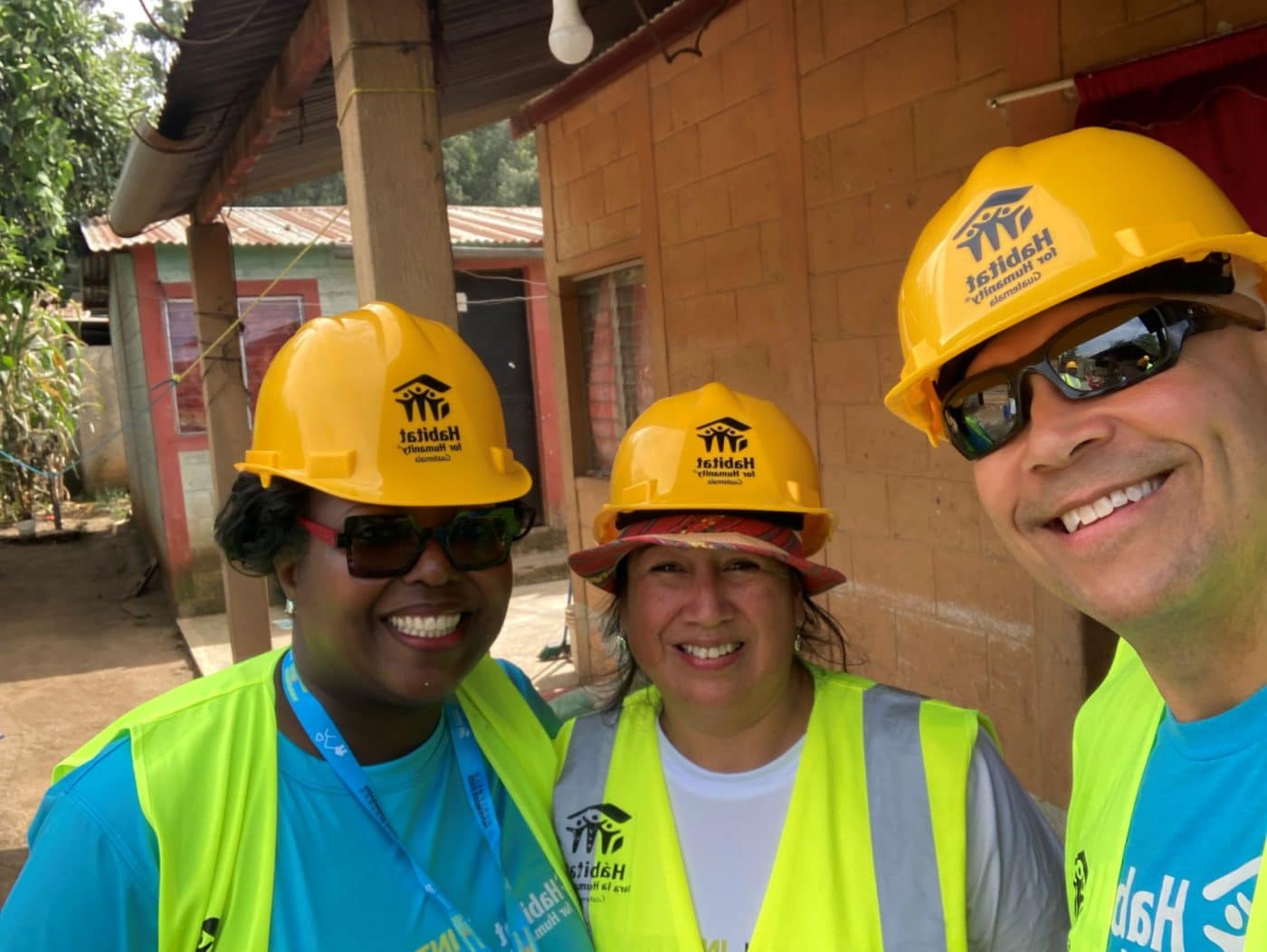 3 people in construction hats and vests smile in front of a construction site