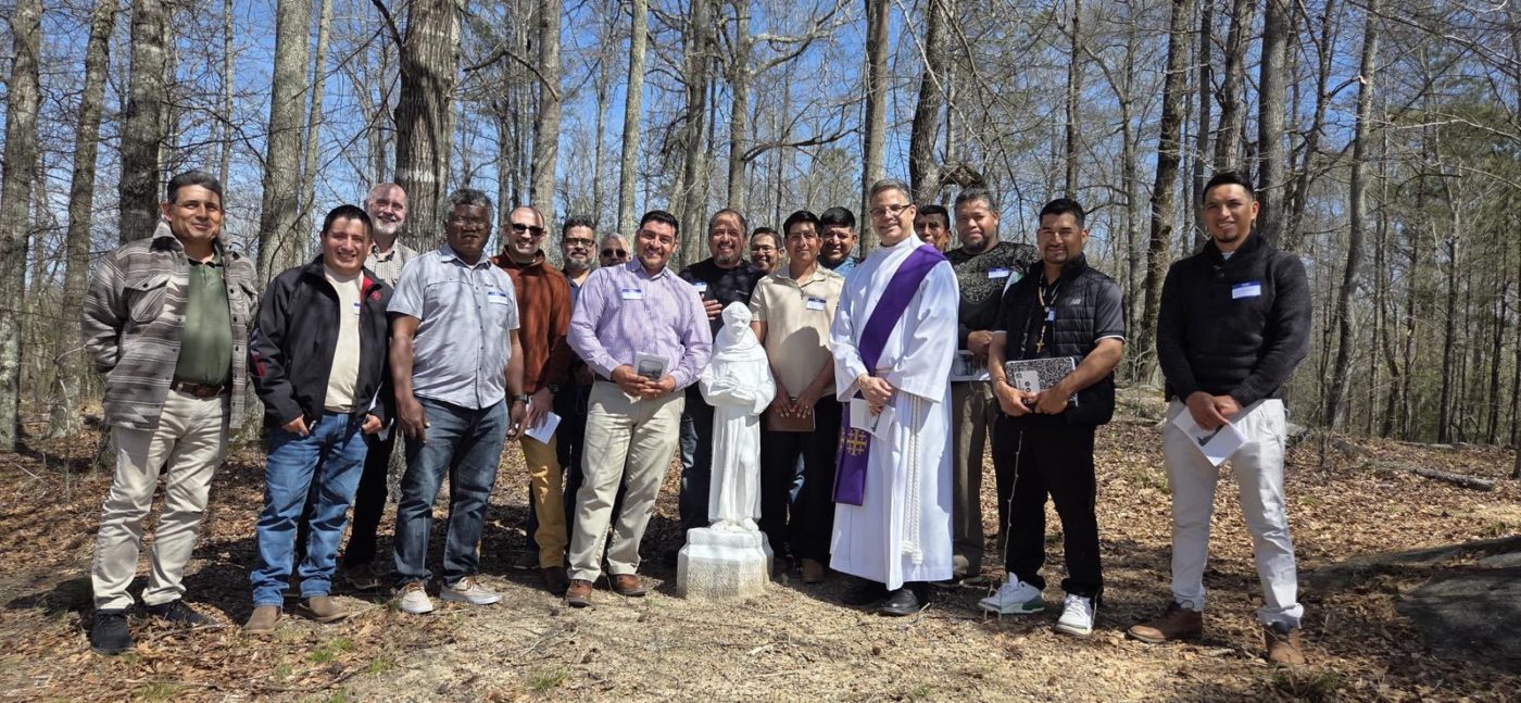 a group of men, including one in clergy attire, pose outside with a statue in the center, trees are in the background