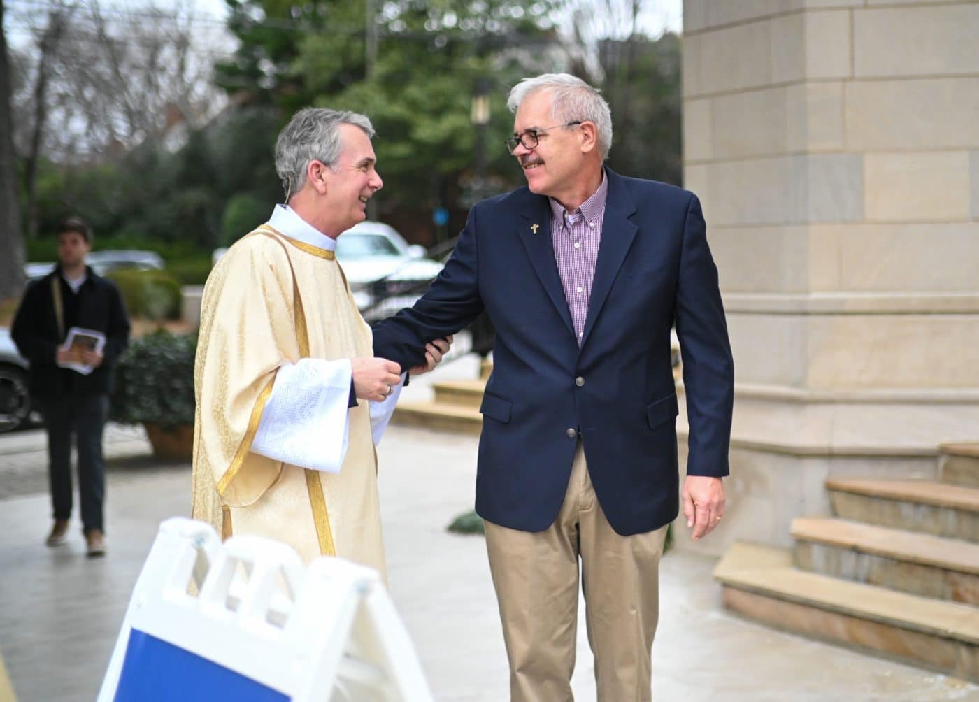 a man in clergy attire greets a man in a suit outside of a church
