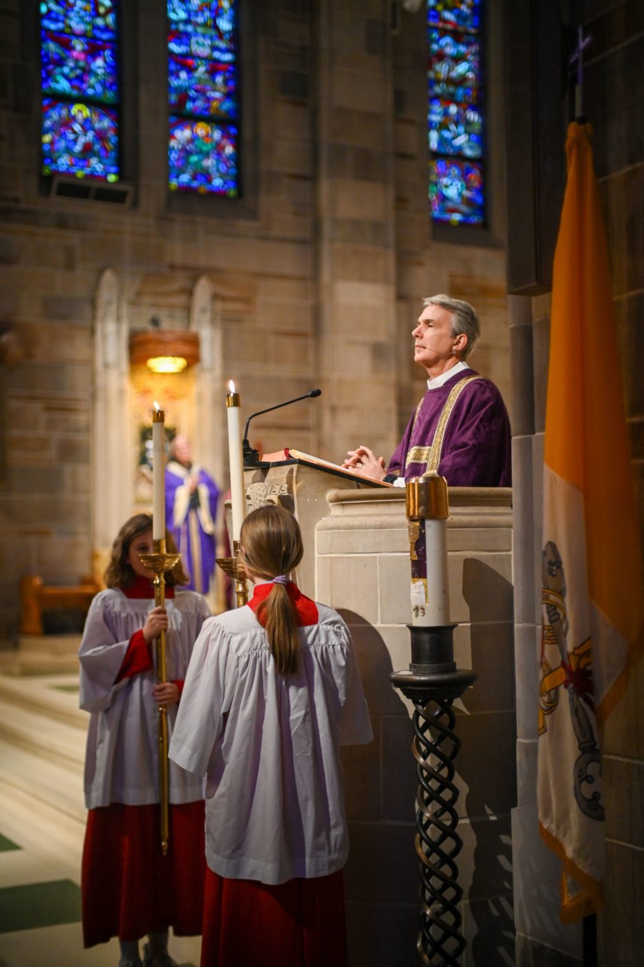 A man in clergy attire stands at a podium with altar servers holding candles on either side in a cathedral with large stain glass windows