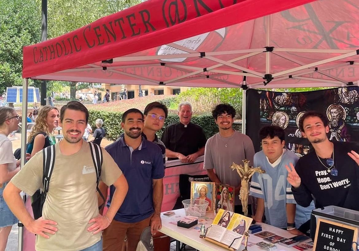 A group of students and one priest pose under a KSU Catholic Center event tent on a busy college campus