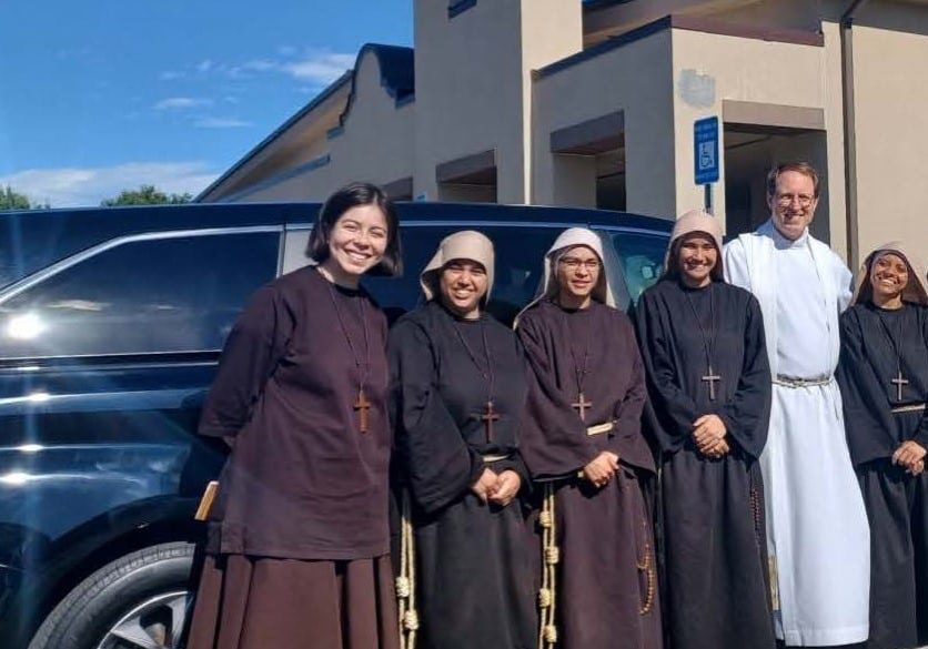 Six women in religious garments and one Catholic priest in religious garments smiling for the camera in front of a vehicle