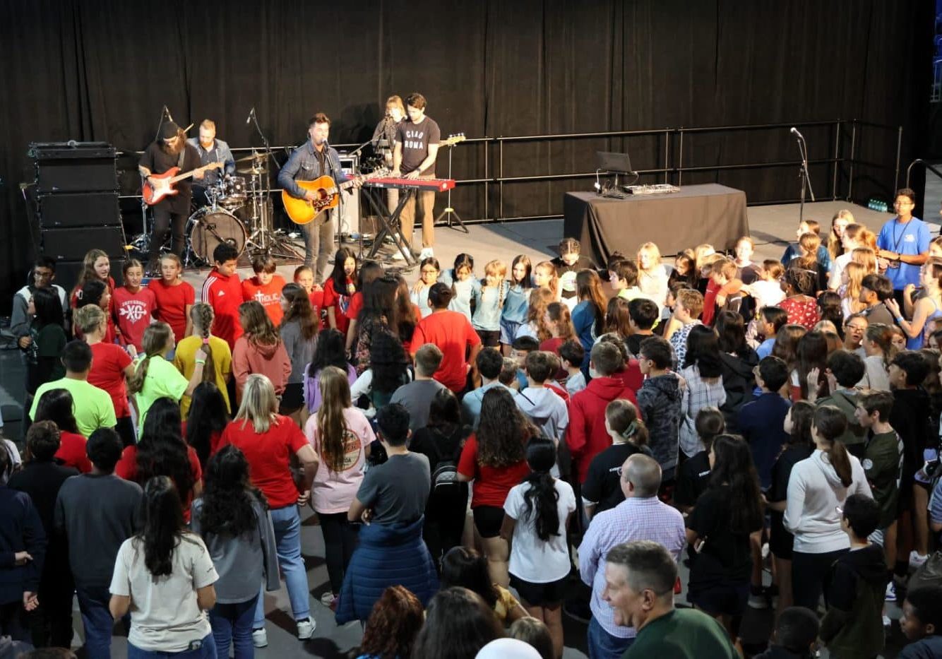 A large group of children stand while watching a band play music on stage