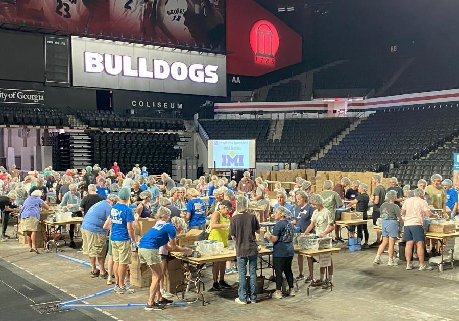 Around 100 people of all ages work to pack meals as they surround various tables scattered throughout a sports arena.