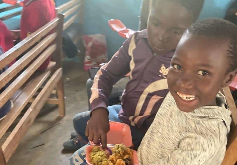 Children sitting on an indoor bench smiling as they share a meal